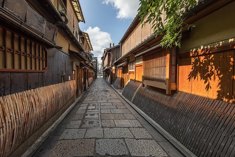 A quiet cobbled street in Gion between wooden houses with bamboo screens, willow leaves catching the afternoon sun.