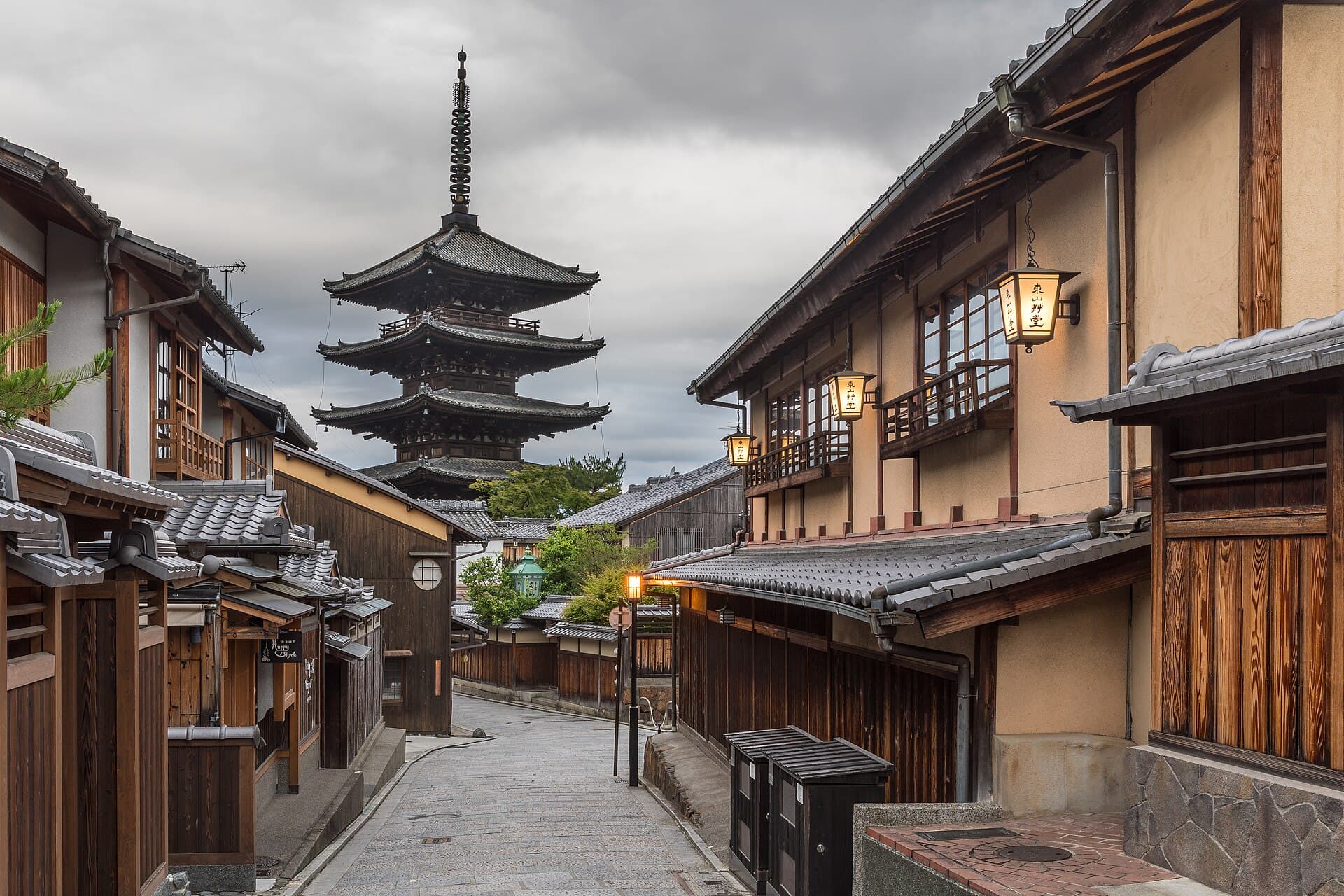 An empty stone lane in Higashiyama at dawn, lit lanterns on wooden houses and the Yasaka pagoda rising under a grey sky.