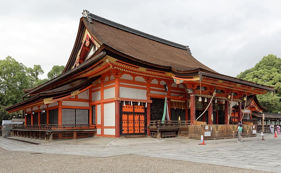 The vermilion and white main hall of Yasaka Shrine under a curved bark roof, with a few visitors on the stone forecourt.